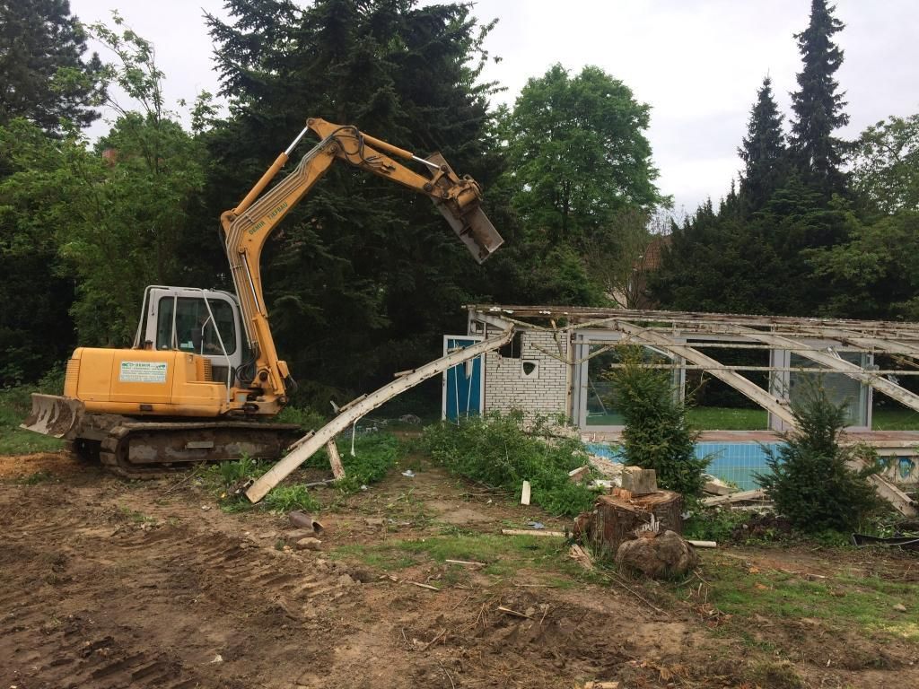Ein gelber Bagger zerstört ein Gebäude neben einem Schwimmbad.