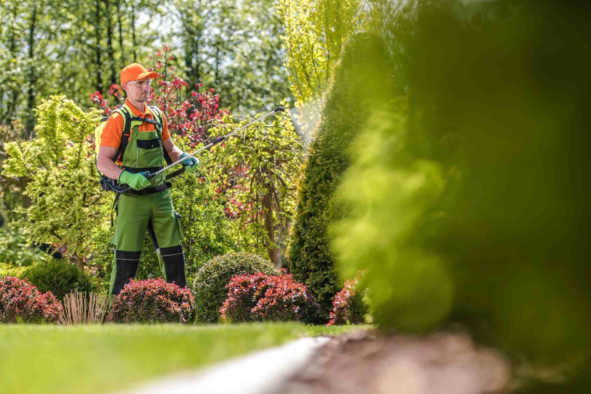 Un jardinero con chaleco naranja se encuentra en un jardín verde exuberante con arbustos y árboles en flor.