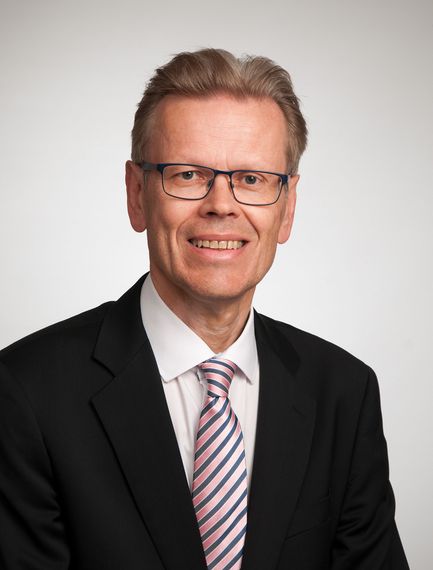 Muotokuva hymyilevästä miehestä mustassa puvussa, valkoisessa paidassa ja raidallisessa vaaleanpunaisessa solmiossa vaalealla taustalla