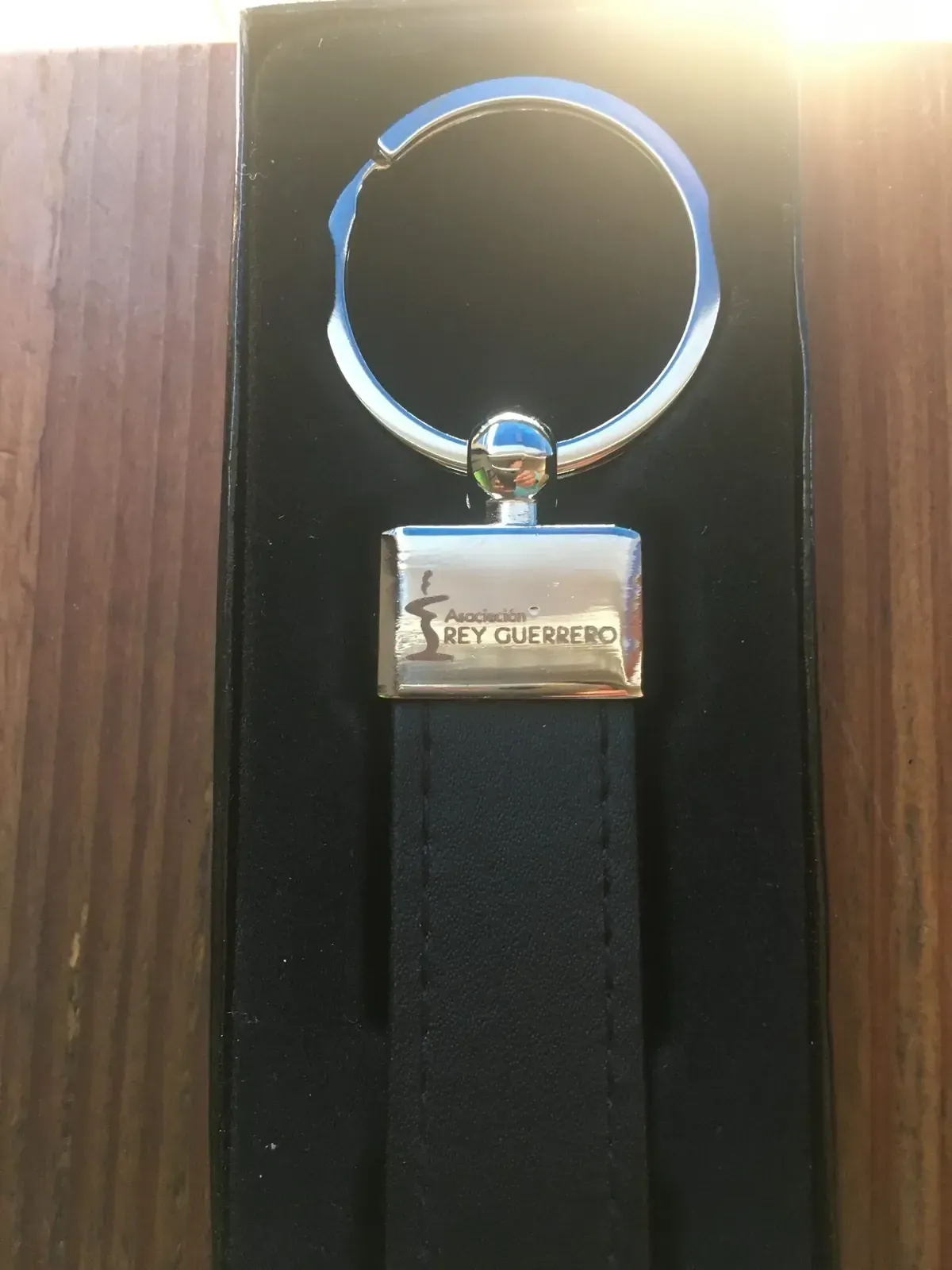 Llavero de cuero negro con llavero y etiqueta plateados, dentro de una caja negra.