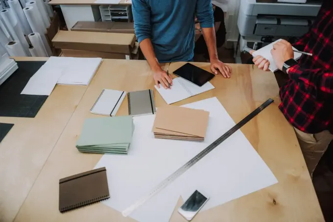 Dos personas en una mesa de madera revisan papeles y libros. También hay papeles de colores y una regla de metal.