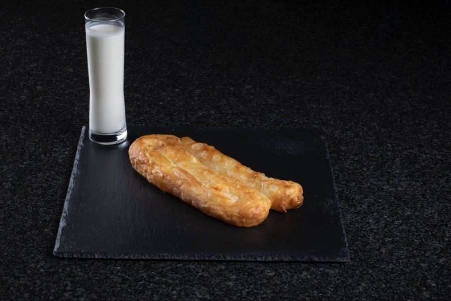 Ein Stück Burek liegt auf einem schwarzen Teller neben einem Glas Milch. Foto von der Burek House GmbH.
