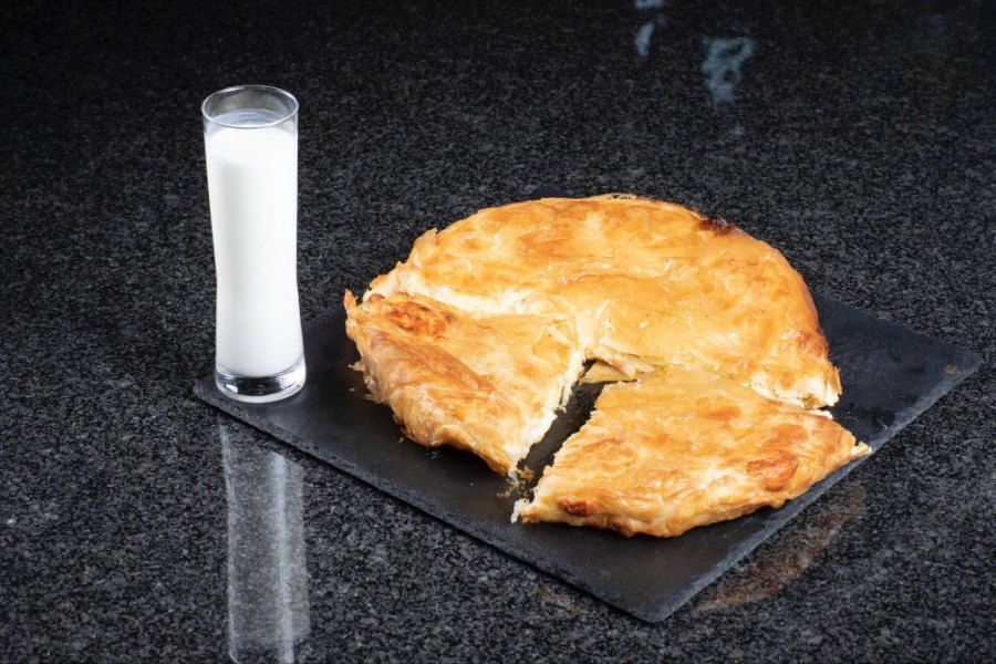 Auf einem schwarzen Teller steht ein Burek neben einem Glas Milch. Foto von der Burek House GmbH.
