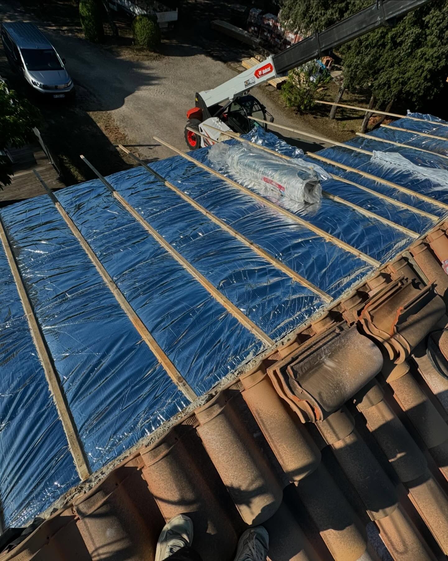 La toiture est en cours de réparation avec de l'isolant bleu et des tuiles ; une grue livre les matériaux.
