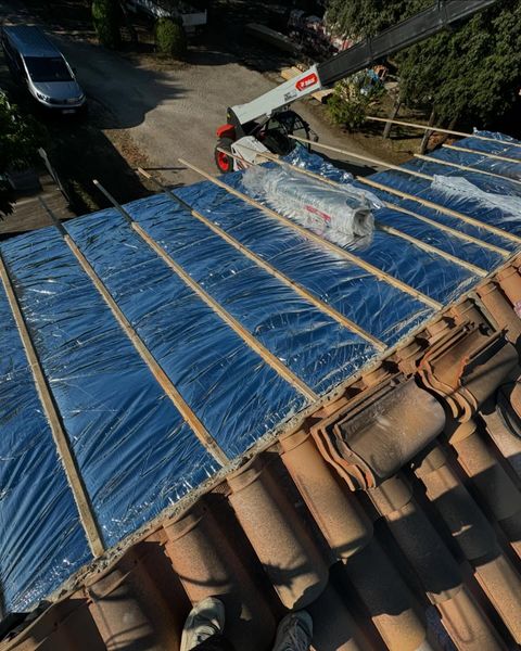 La toiture est en cours de réparation avec de l'isolant bleu et des tuiles ; une grue livre les matériaux.