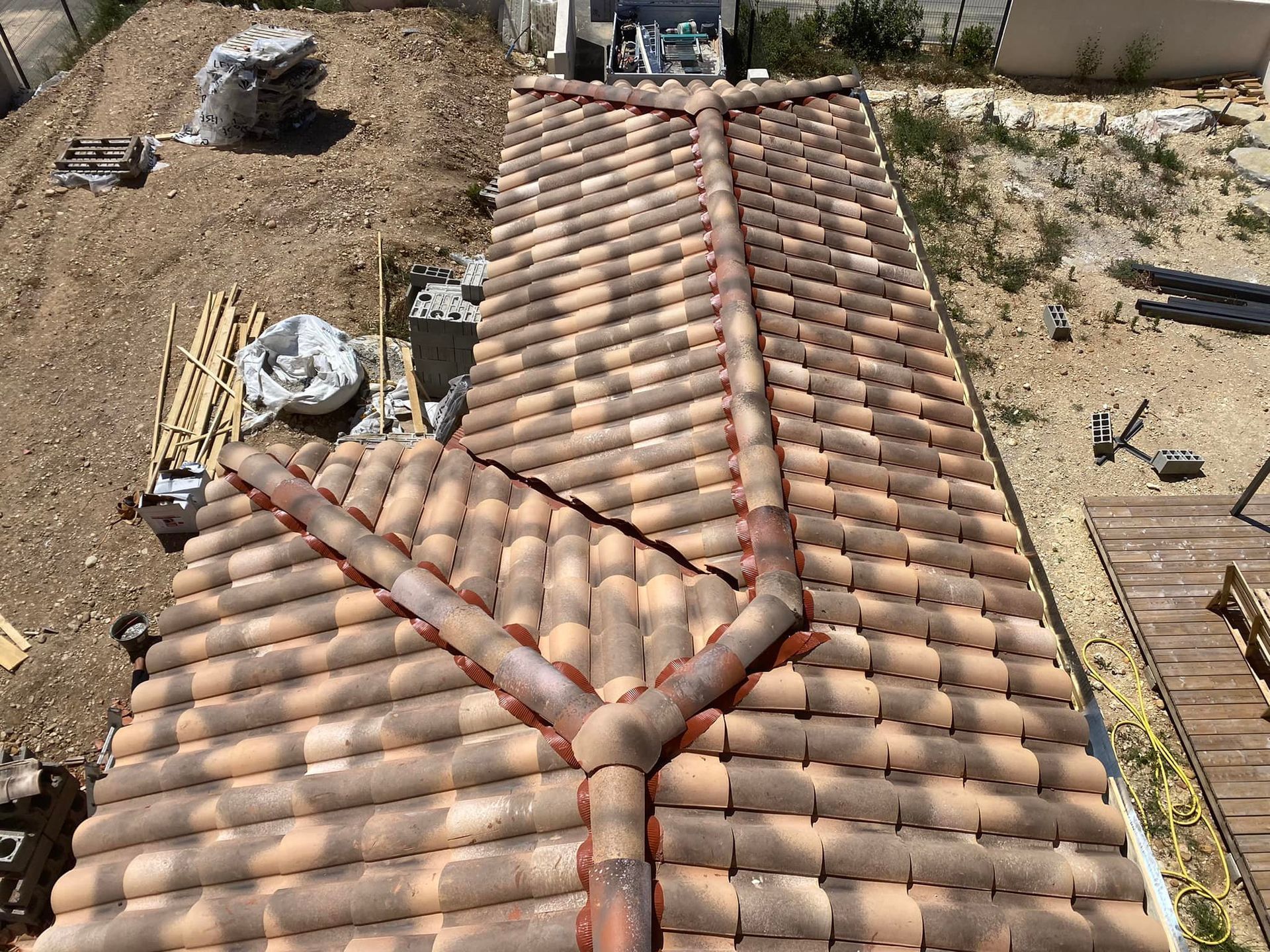 Vue aérienne d'un toit de tuiles aux tons terreux avec une intersection de lignes de faîtage, situé à proximité d'un chantier de construction.