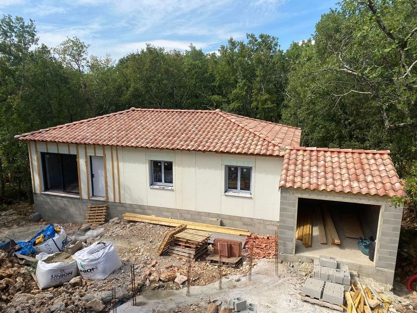Maison en construction avec toit de tuiles rouges, entourée d'arbres.