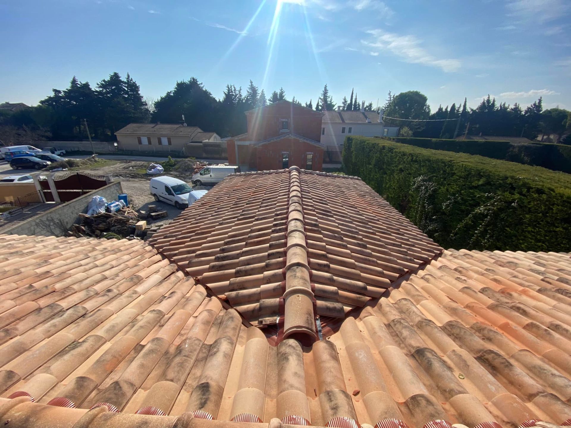 Toit de tuiles en terre cuite à motifs arqués, soleil, arbres et bâtiments en arrière-plan.