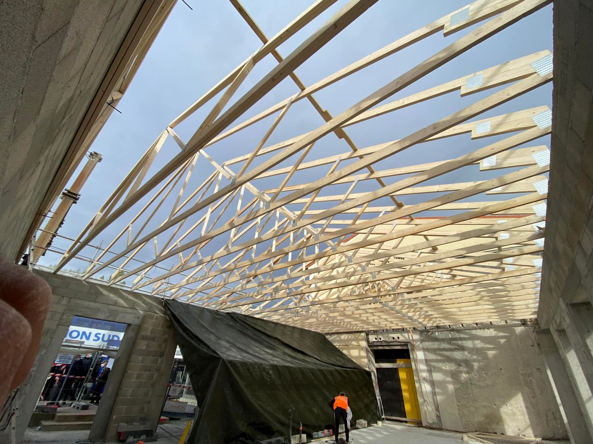Intérieur d'un chantier de construction montrant des fermes de toit en bois au-dessus de murs en blocs de béton et une zone couverte avec une personne à proximité.
