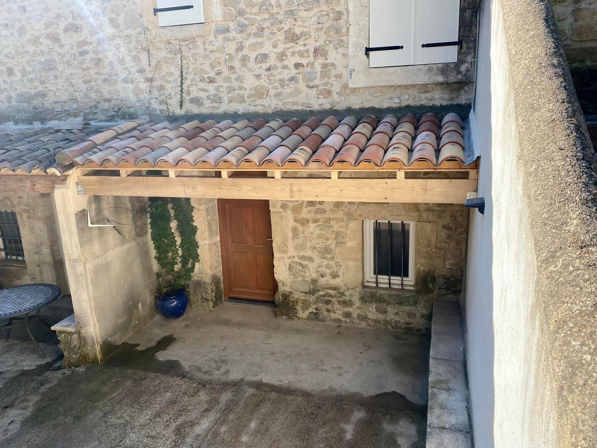Entrée couverte avec murs en pierre, porte brune, fenêtre à barreaux et toit de tuiles.