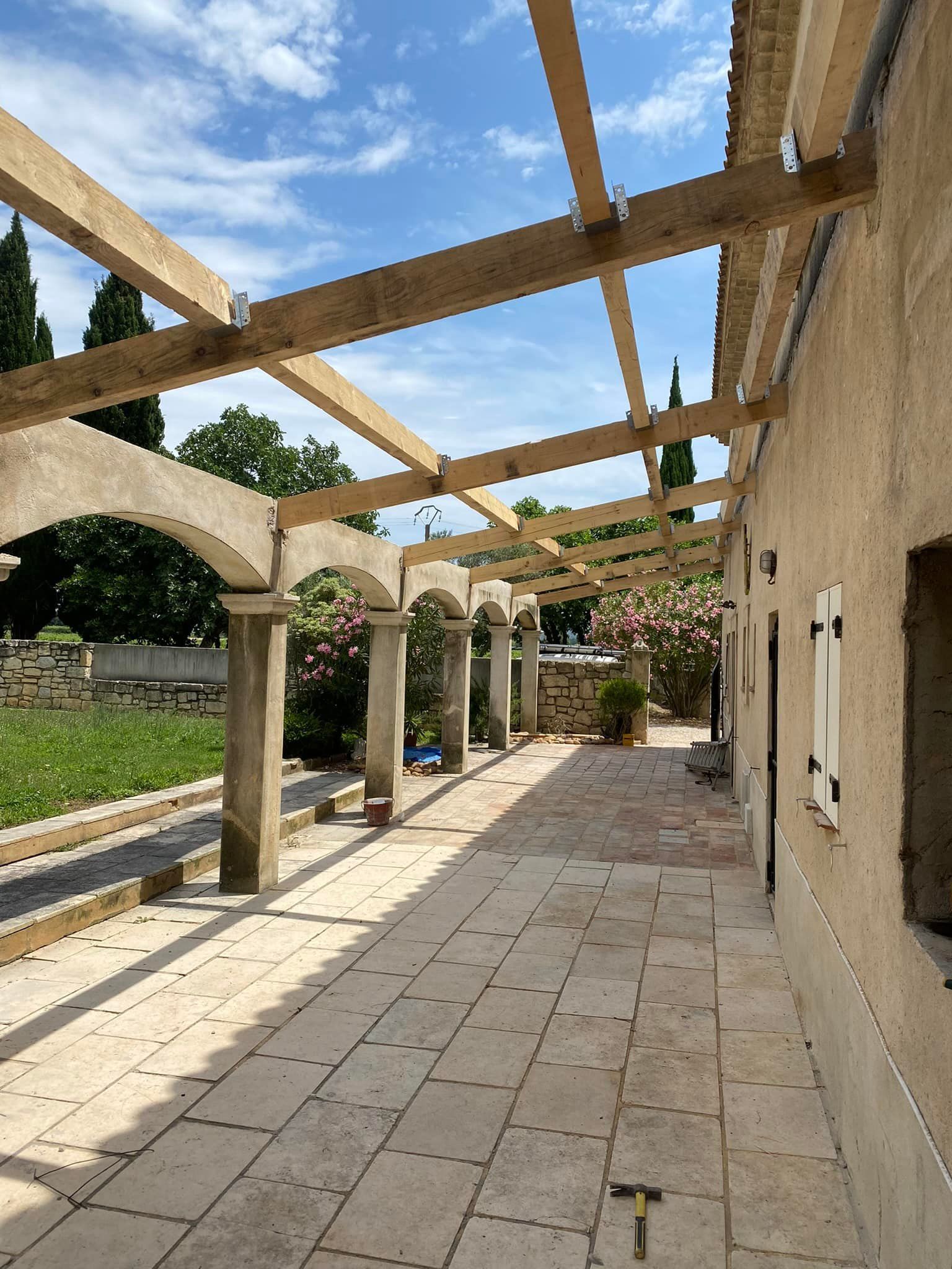 Construction d'une pergola au-dessus d'un patio carrelé, à côté d'un bâtiment aux colonnes de pierre, sous un ciel bleu.