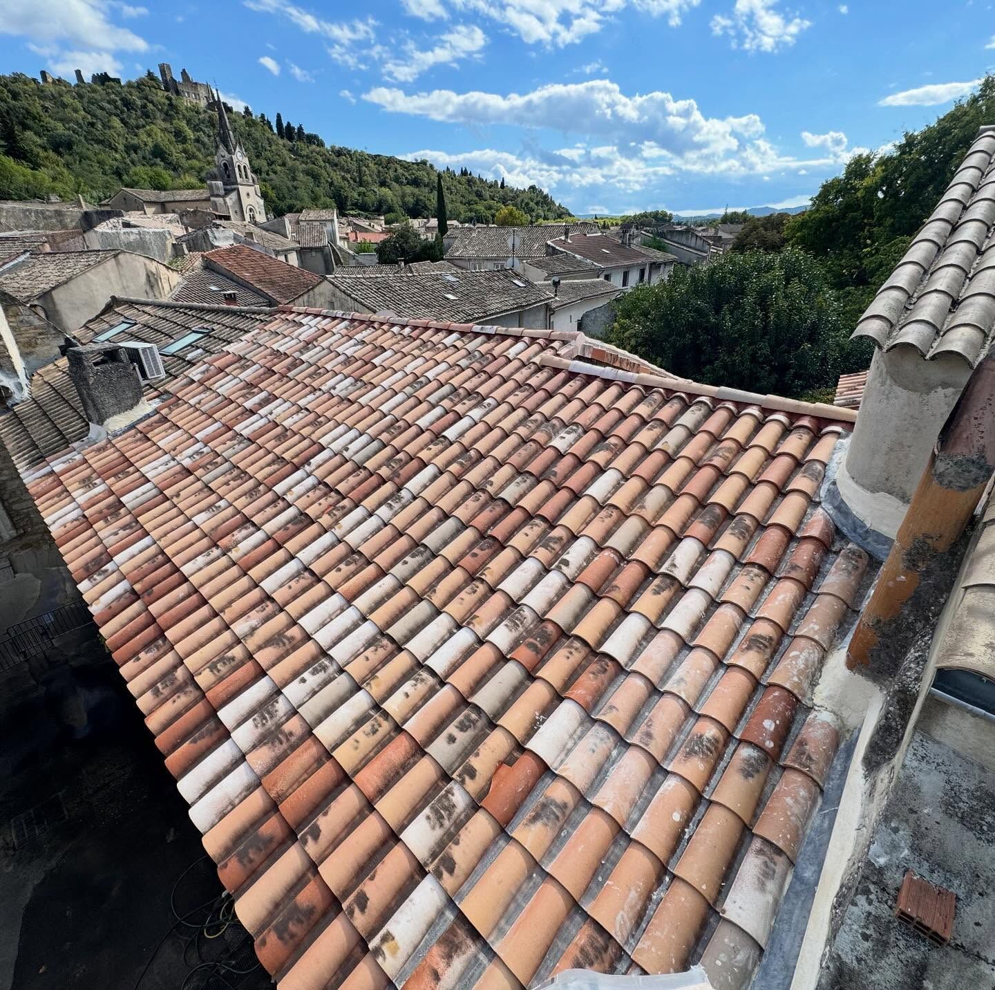 Vue en plongée sur un toit de tuiles en terre cuite patiné par le temps, dans un village européen avec une colline en arrière-plan.