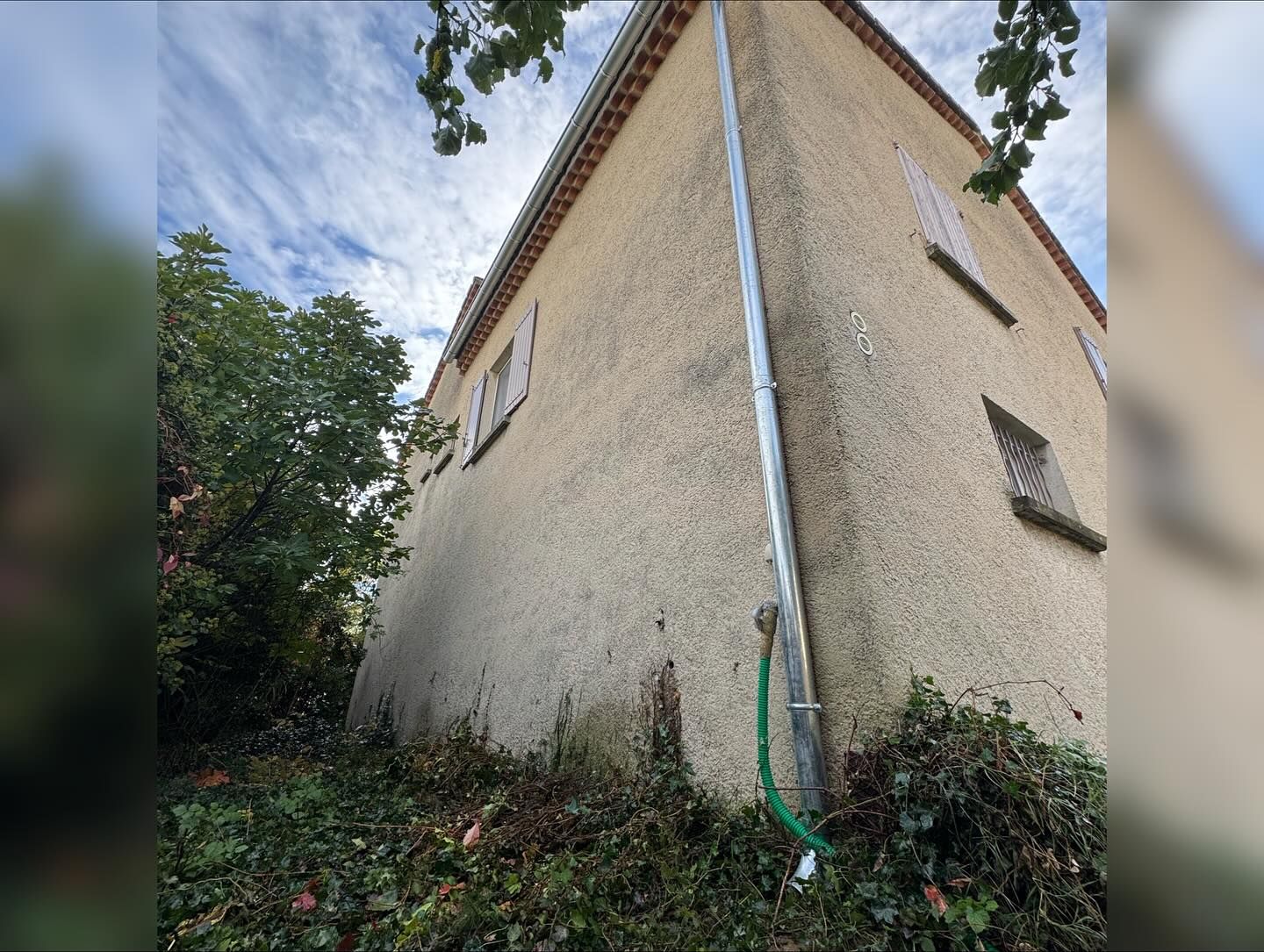 Bâtiment d'angle en stuc beige avec une descente de gouttière, entouré de verdure. Ciel nuageux au-dessus.