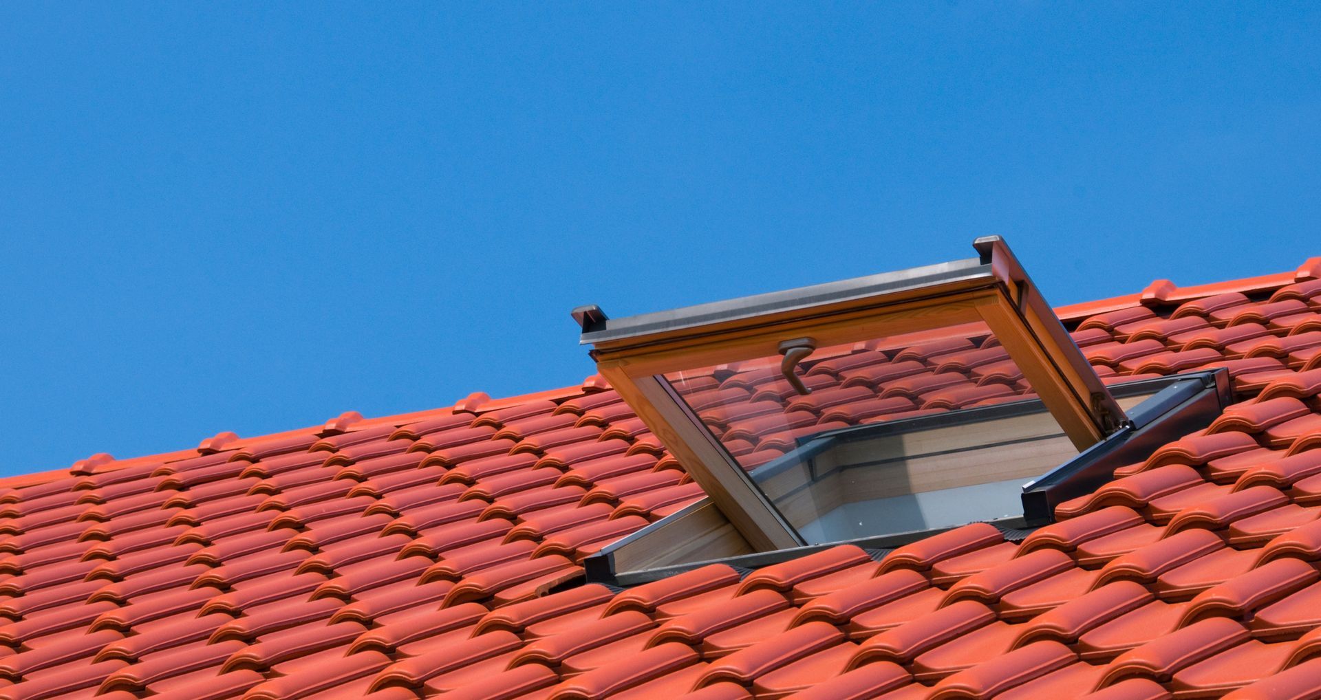 Une fenêtre de toit ouverte, encastrée dans un toit de tuiles rouge vif, se détachant sur un ciel bleu limpide.