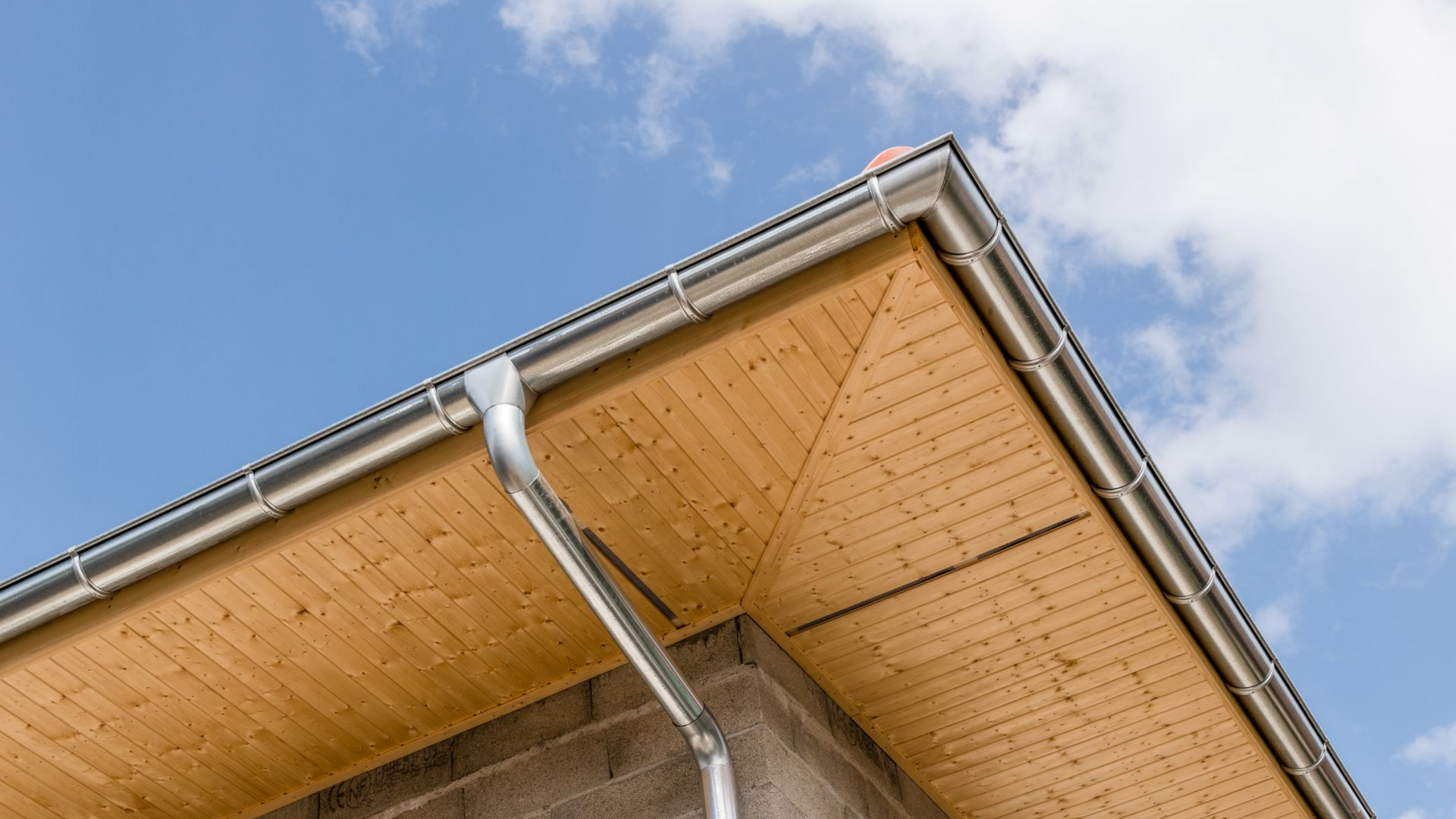 Vue en contreplongée d'un avant-toit en bois avec gouttières métalliques et descente pluviale, sur fond de ciel bleu légèrement nuageux.