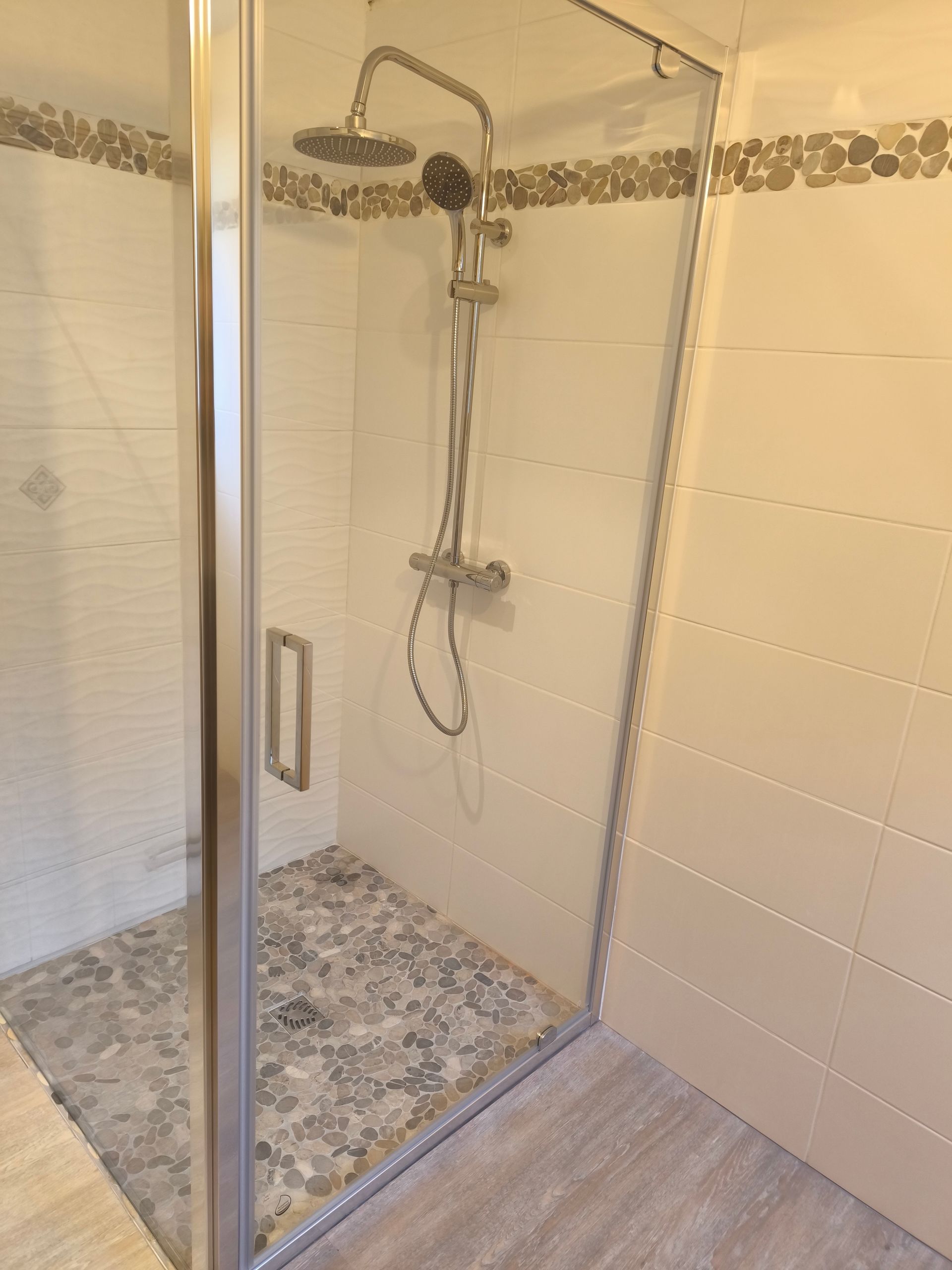Cabine de douche avec porte en verre, sol en galets, murs carrelés blancs et robinetterie argentée
