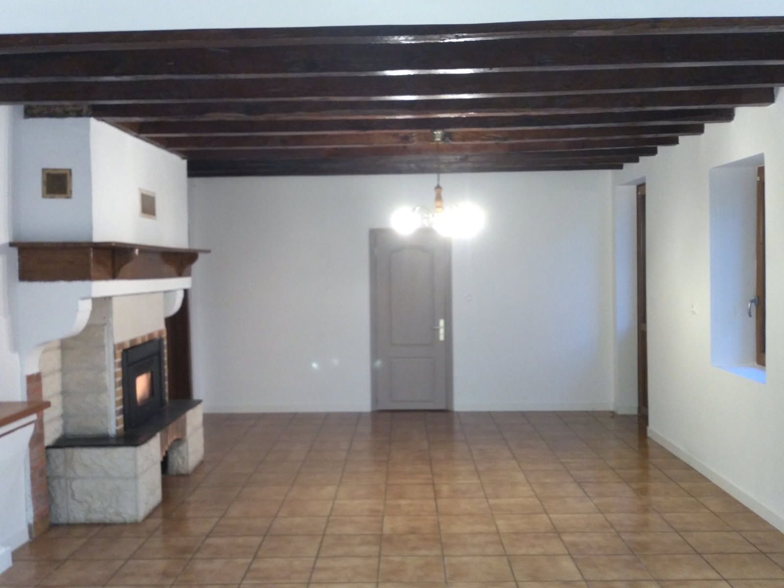 Salon vide avec cheminée, plafond à poutres apparentes, sol carrelé.