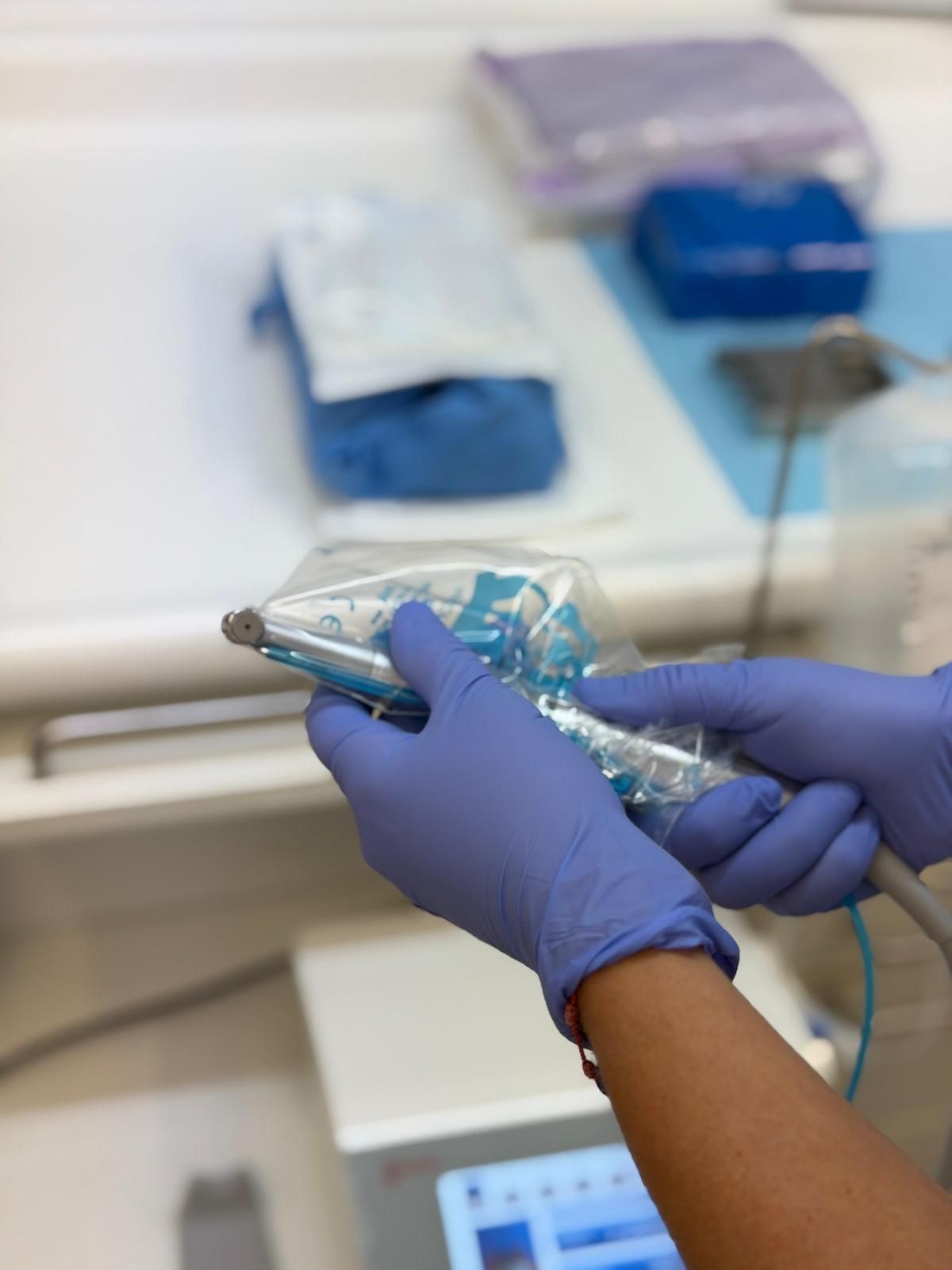 Manos con guantes azules sujetando un taladro dental con funda protectora. El entorno es un espacio de trabajo médico.