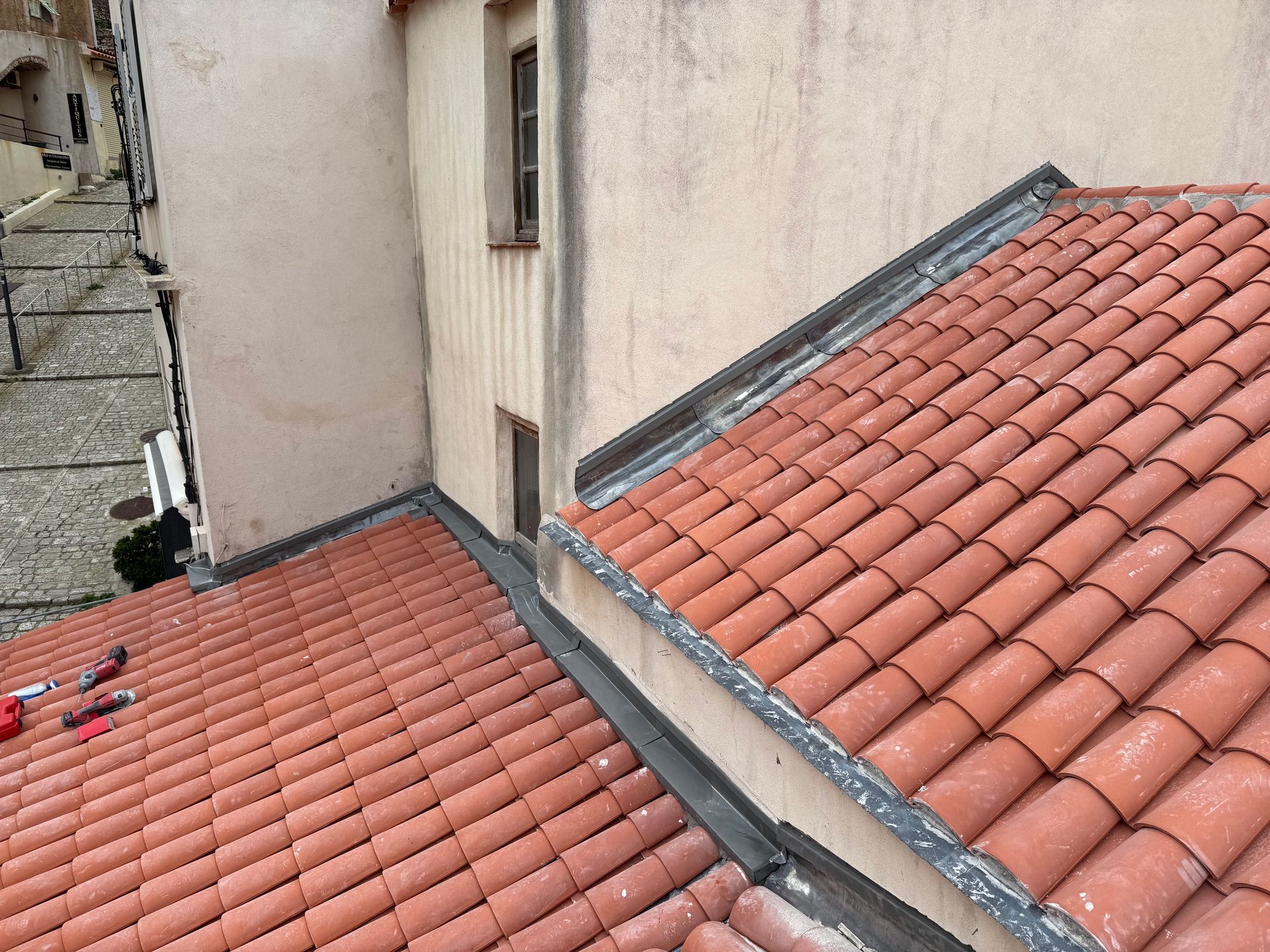 Toits de tuiles en terre cuite rouge jouxtant un bâtiment beige et donnant sur une ruelle pavée.