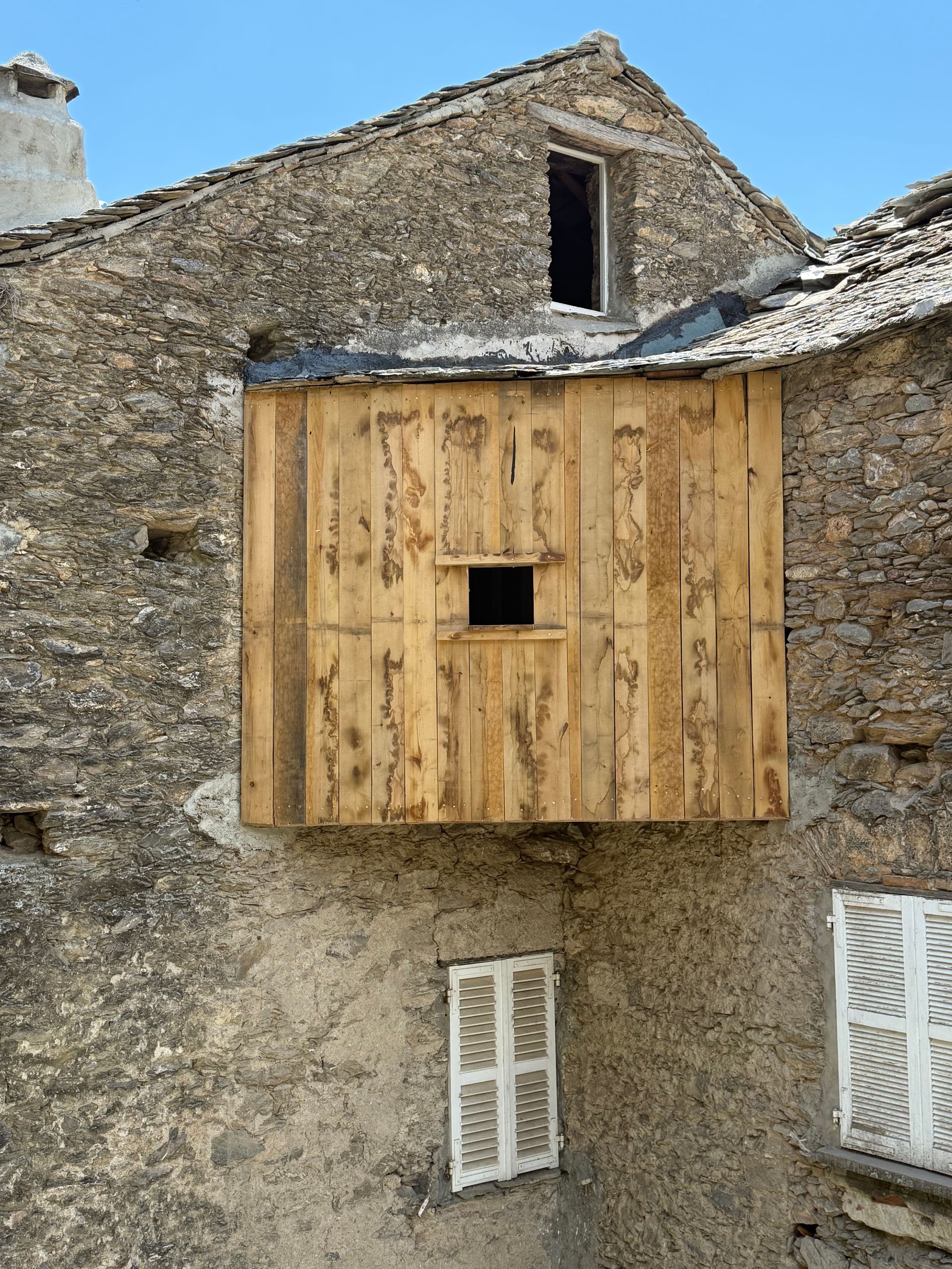 Bâtiment rustique en pierre avec un petit balcon en bois et des fenêtres à volets sous un ciel bleu
