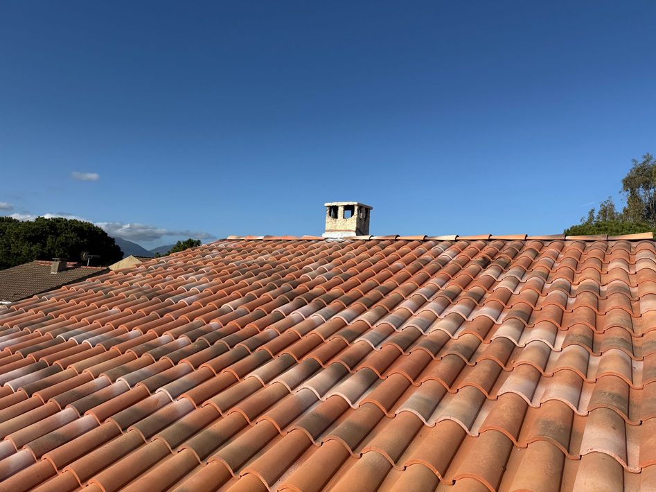 Toit de tuiles en terre cuite rouge sous un ciel bleu limpide, avec une petite cheminée au loin.