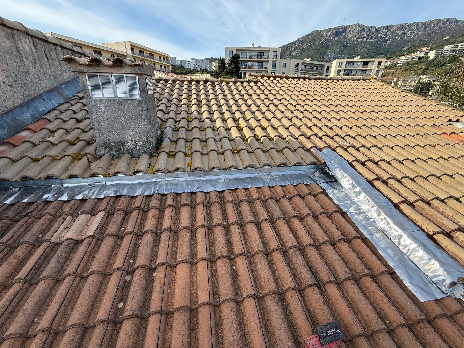 Toits de tuiles en terre cuite avec cheminée, se détachant sur les collines et les bâtiments sous un ciel dégagé.