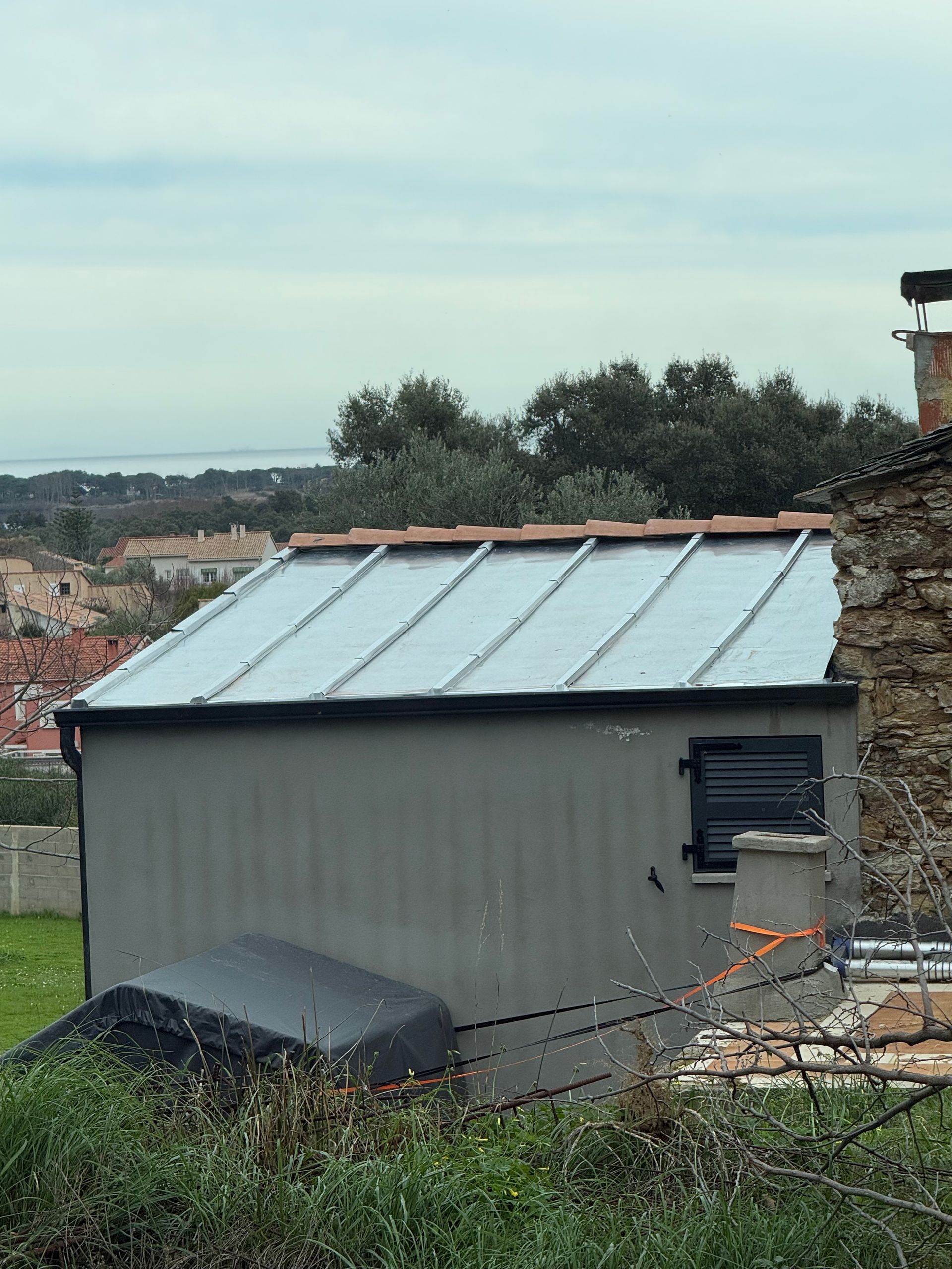 Petit hangar gris au toit en métal léger, situé en zone rurale, avec de la végétation et des maisons en arrière-plan.