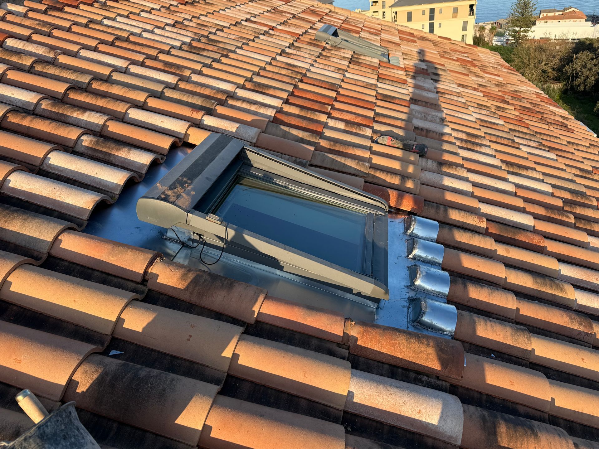 Puits de lumière sur le toit, avec un cadre noir et partie métallique, intégré à une toiture en tuiles de terre cuite orange.