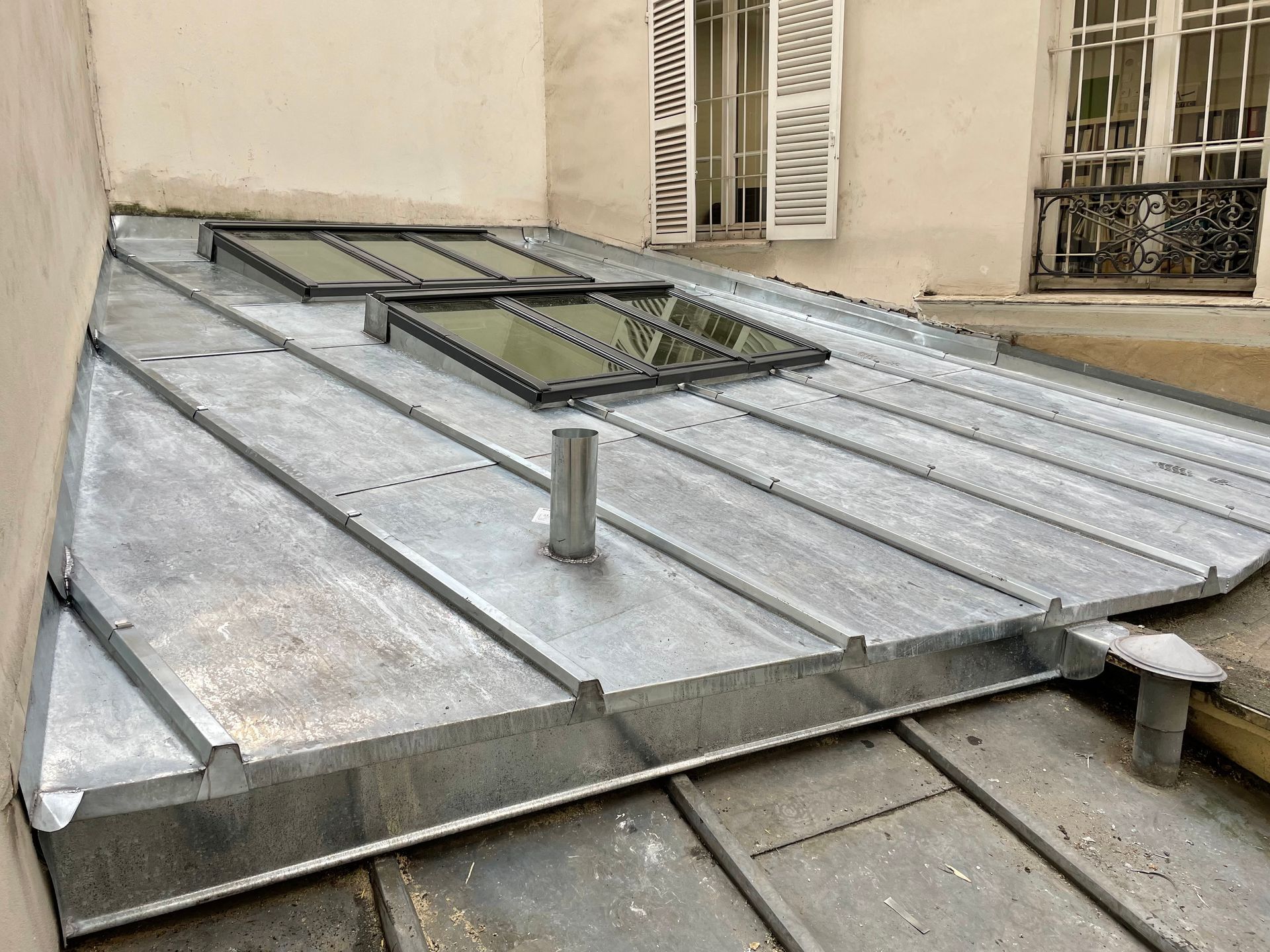 Puits de lumière sur le toit métallique, avec deux fenêtres ouvrantes, jouxtant un mur beige du bâtiment.