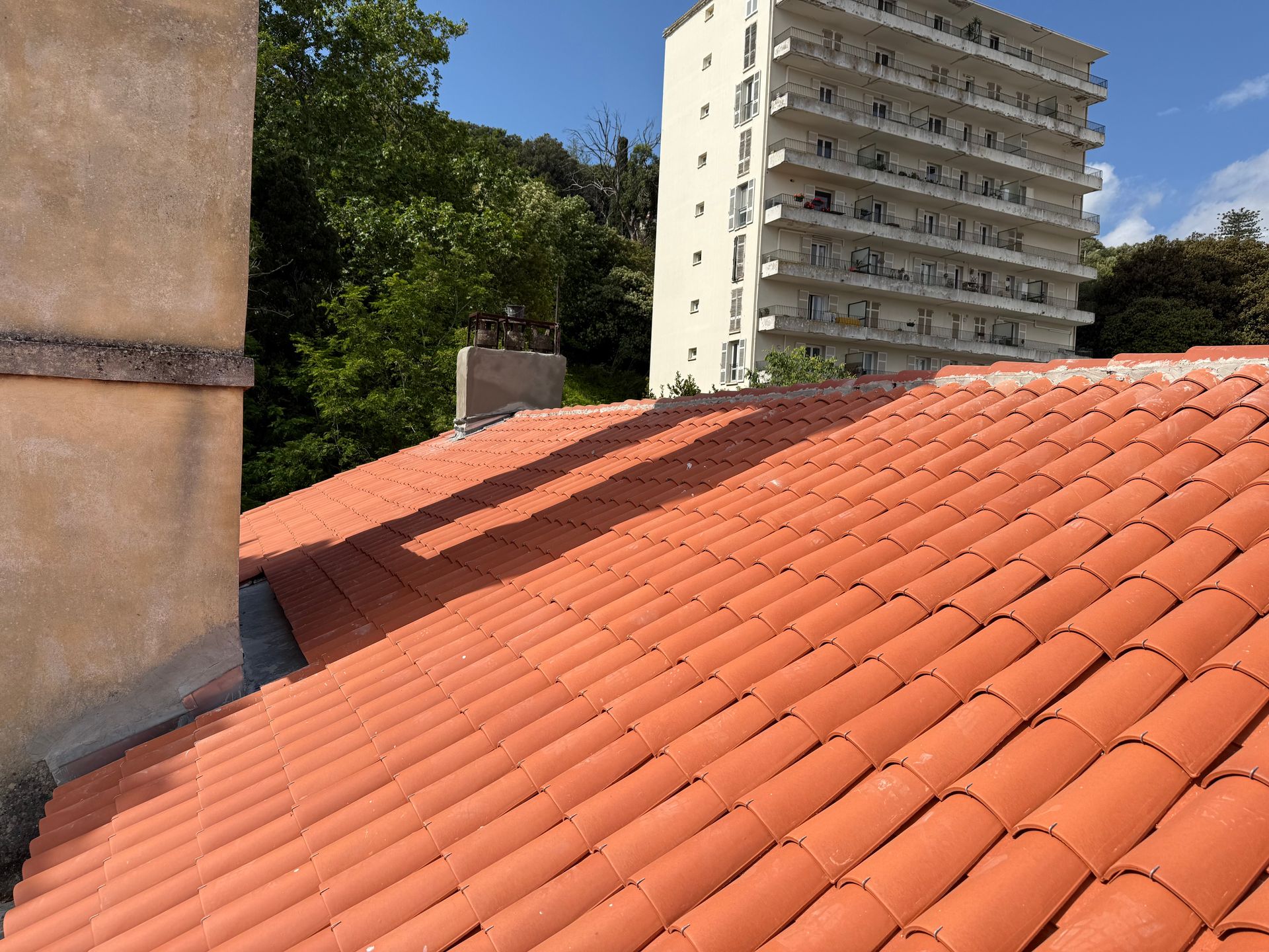 Toiture en tuiles rouges jouxtant un mur beige, avec des arbres et un grand immeuble d'appartements en arrière-plan.