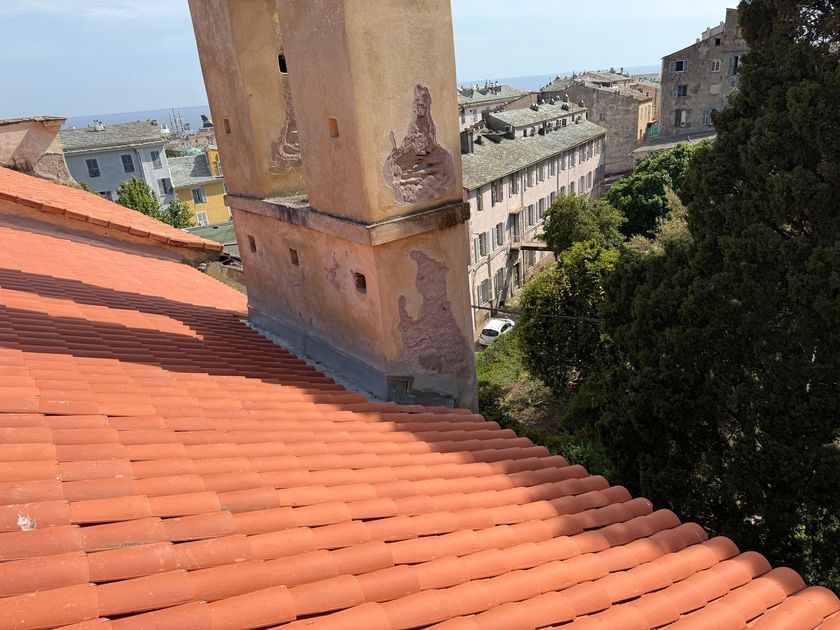 Vue depuis le toit avec des tuiles rouges, une cheminée beige, des arbres et des bâtiments près de la mer