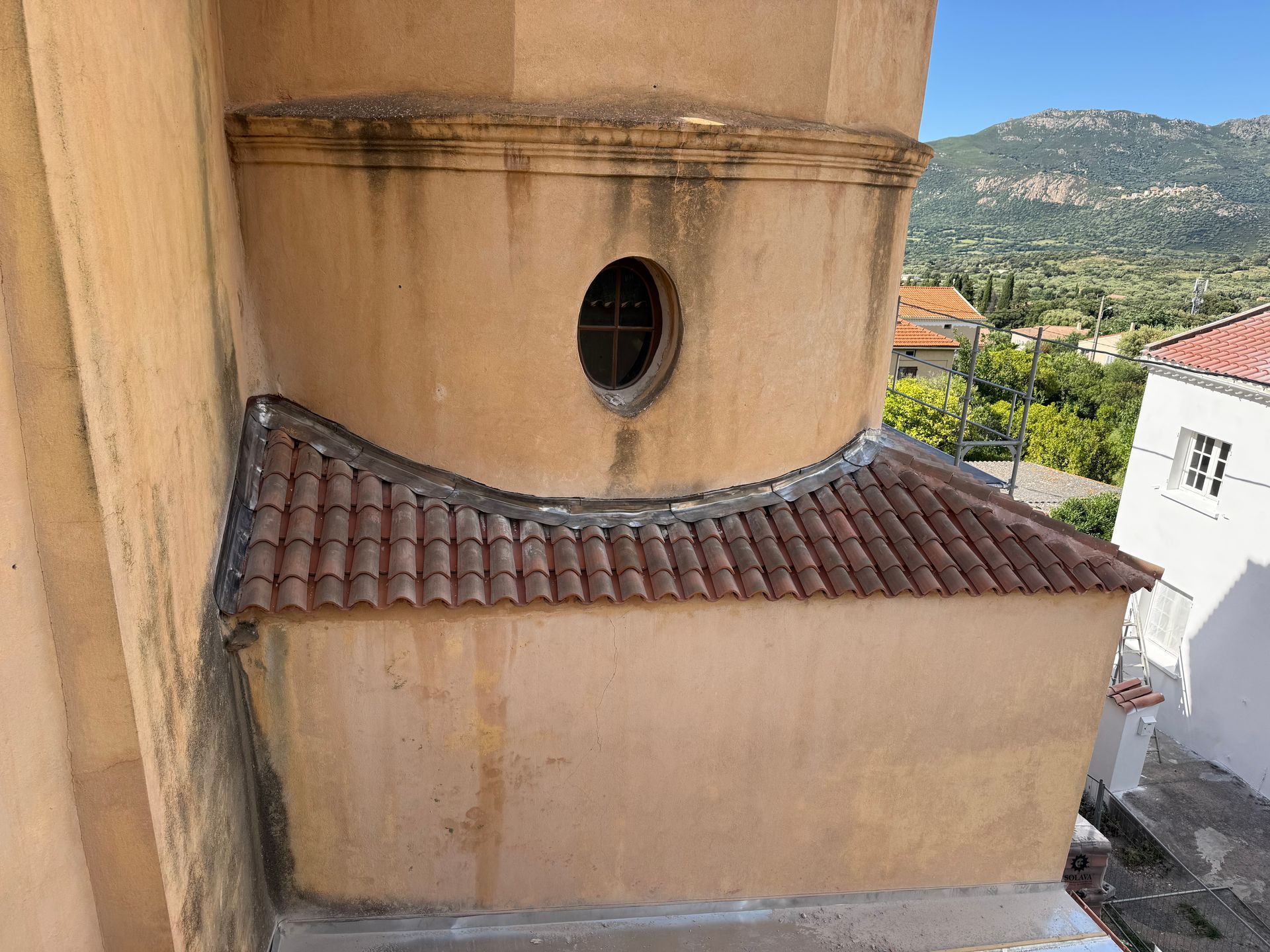 Bâtiment en stuc beige avec une fenêtre ronde et un toit de tuiles rouges, se détachant sur un fond de montagne.