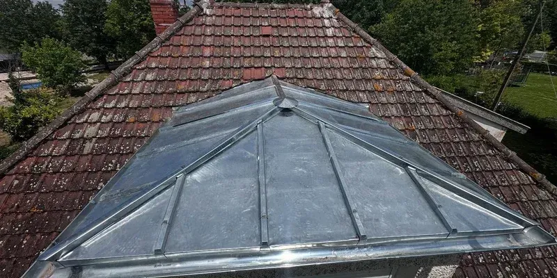 Toit triangulaire en métal sur une maison, entourée de tuiles en terre cuite rouge, avec des arbres en arrière-plan
