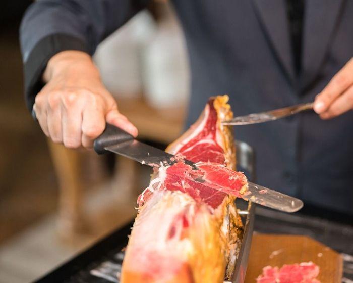 Persona trinchando un jamón curado rojo con un cuchillo en un bufé.