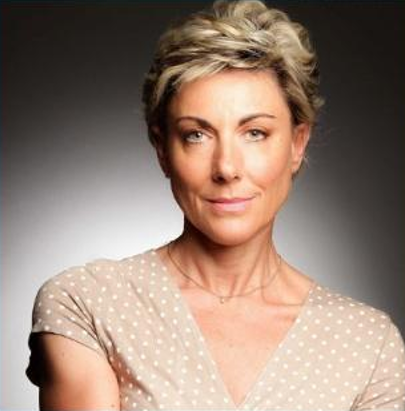 Femme blonde aux cheveux courts, esquissant un sourire, vêtue d'un haut à pois