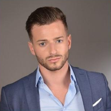 Homme aux cheveux bruns courts et coiffés, barbe, veste de costume bleue et chemise à col bleu clair