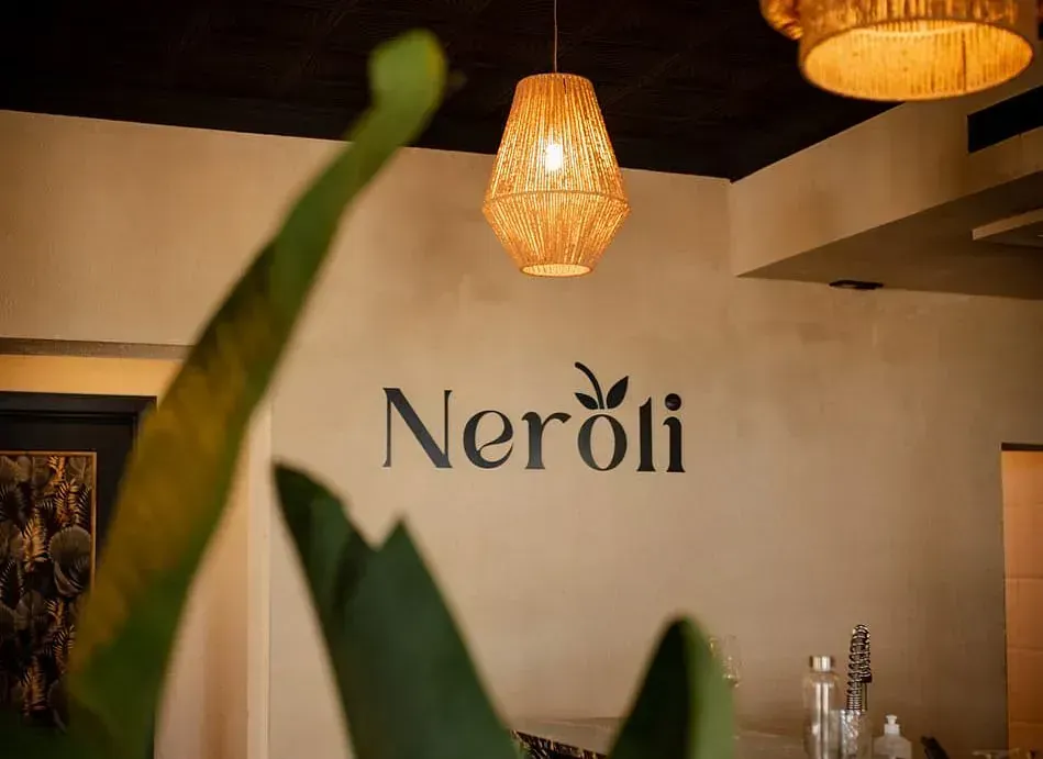 Logotipo "Neroli" en una pared de color claro, bajo una lámpara colgante de mimbre. Planta en primer plano, interior.