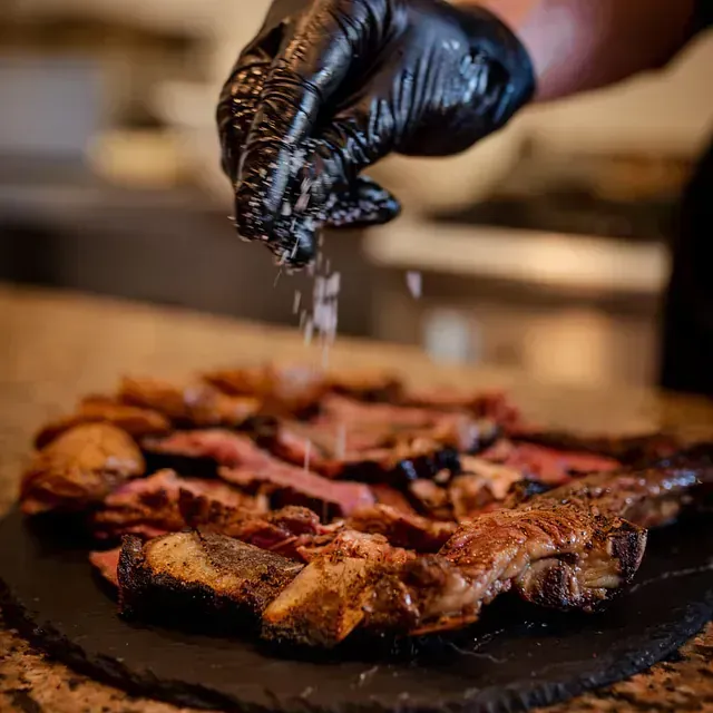Persona con guantes negros salando carne asada en rodajas sobre una tabla de pizarra.