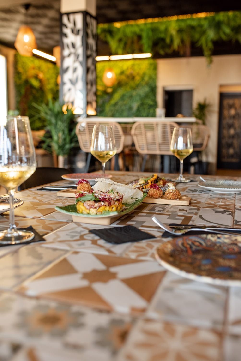 Mesa de restaurante puesta con comida, copas de vino y una colorida superficie de azulejos. Pared verde exuberante al fondo.