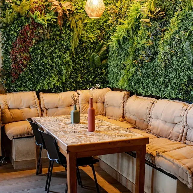 Acogedor reservado con cojines color canela y pared vegetal verde, mesa de madera con botellas.