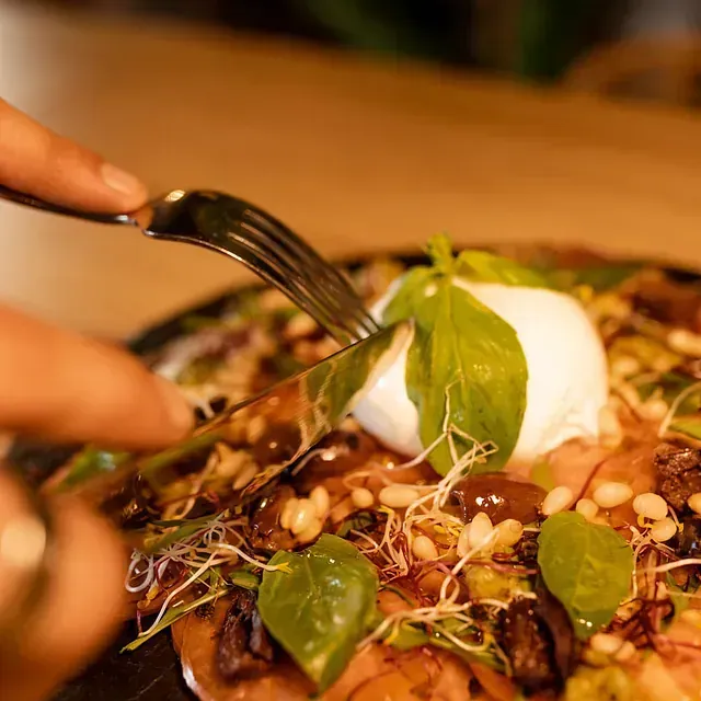 Cortando con tenedor y cuchillo una pizza cubierta con una bola de queso blanco, albahaca y brotes.