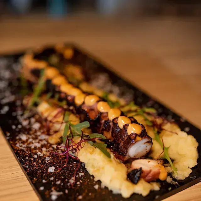 Pulpo a la plancha sobre puré de patatas, rociado con salsa, adornado con hierbas en un plato negro.