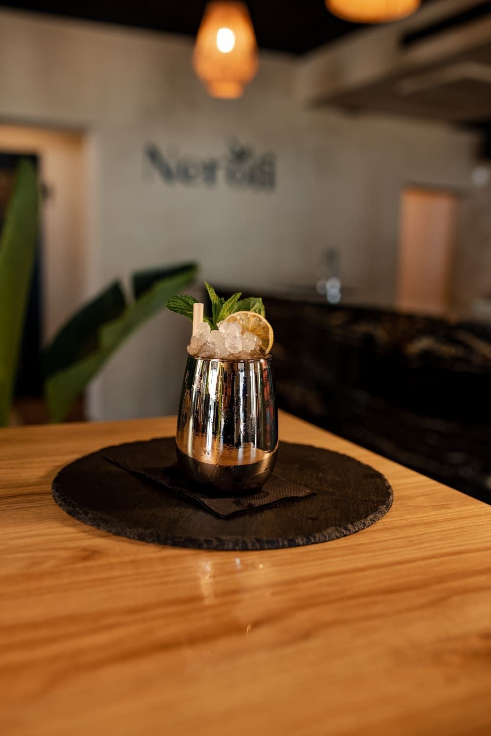 Cóctel servido en una copa de plata, adornado con menta y fruta, sobre una mesa de madera.