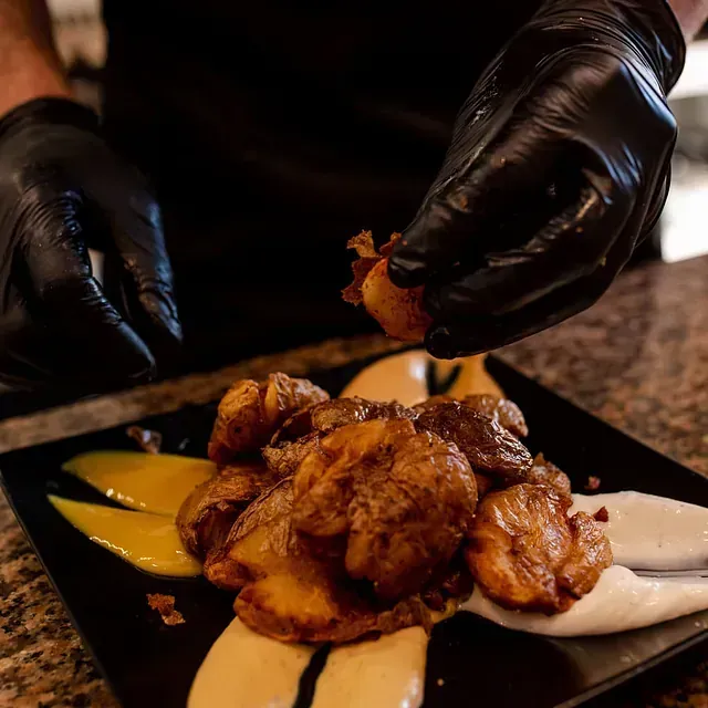 Manos enguantadas de negro colocan patatas asadas con salsas en un plato negro.
