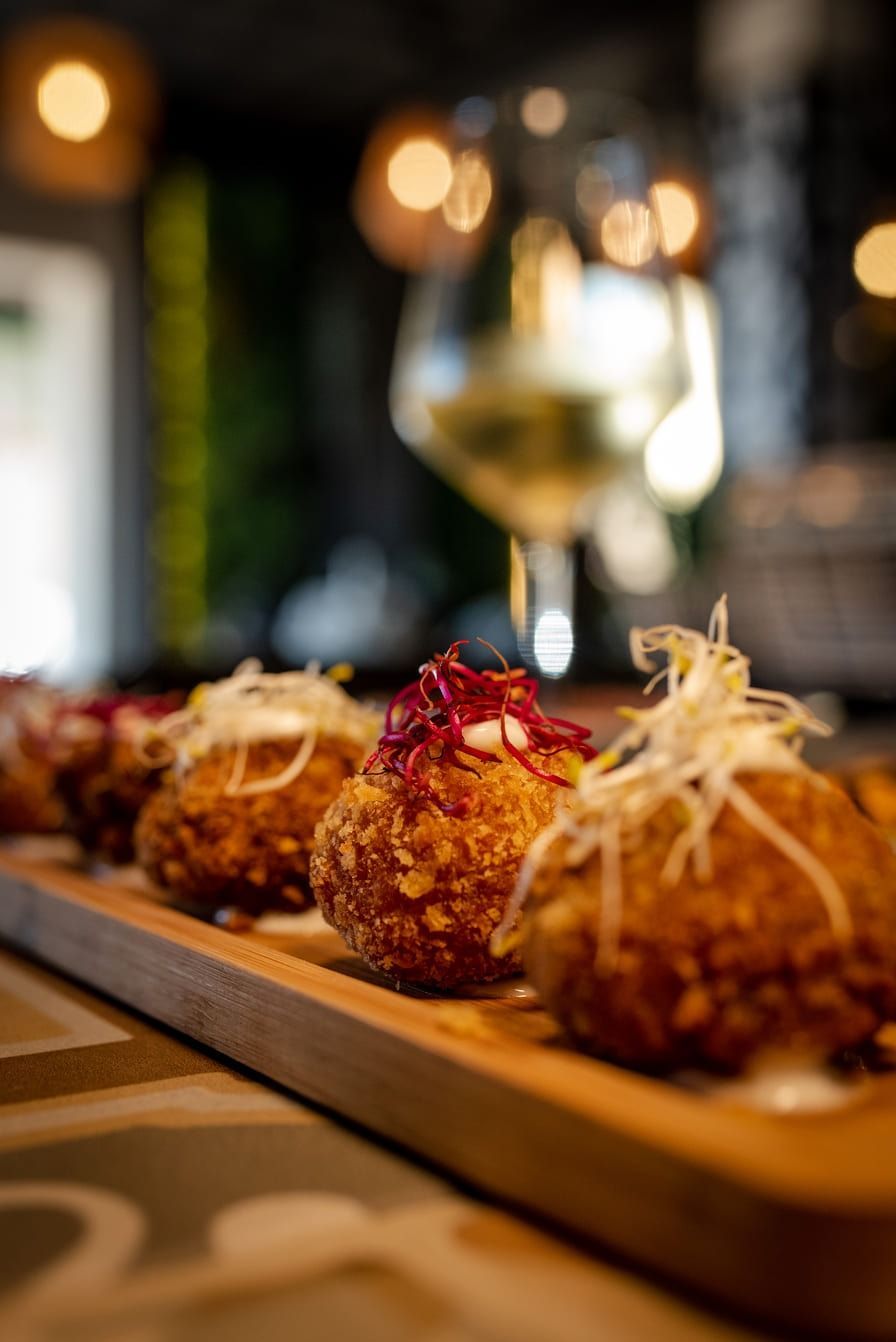 Aperitivos fritos en bandeja de madera, con una copa de vino desenfocada al fondo.
