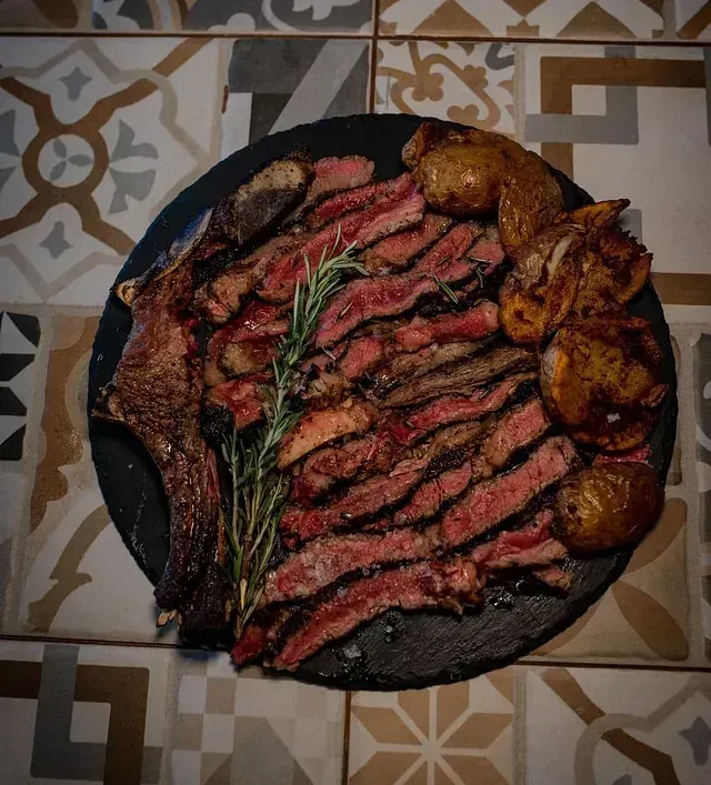Filete de ternera en rodajas con patatas y romero en un plato negro, sobre un fondo de azulejos estampados.