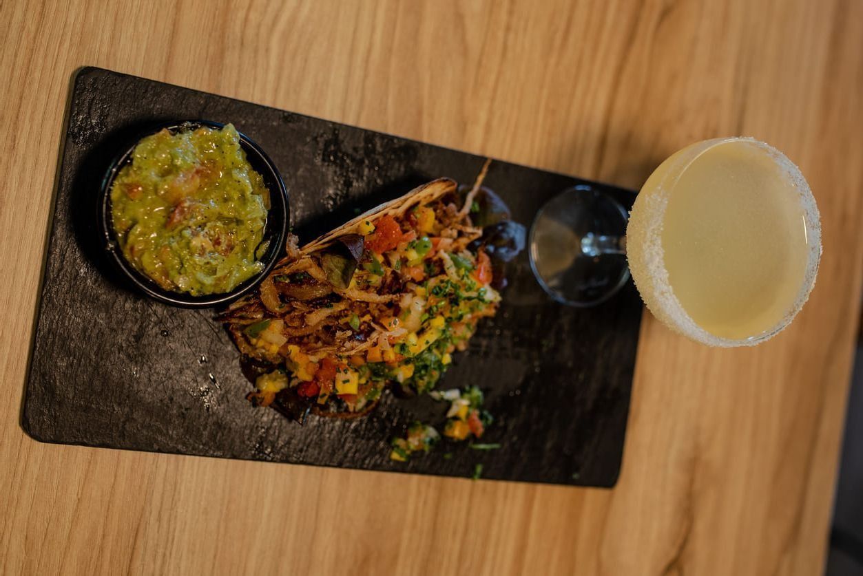 Taco y guacamole en una tabla de pizarra, con una bebida en una copa de tallo sobre una mesa de madera.