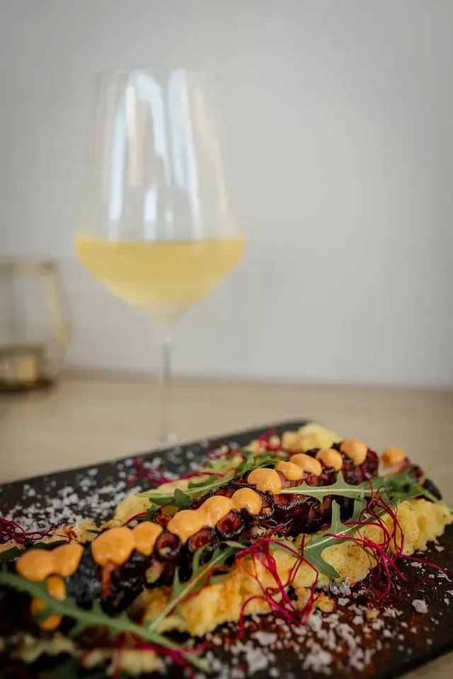 Plato de pulpo a la plancha con salsa, verduras y una copa de vino blanco.