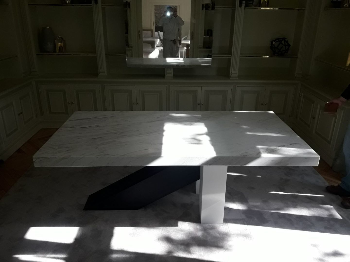 Mesa con tapa de mármol y base en blanco y negro delante de estantes empotrados; una persona se refleja en un espejo.