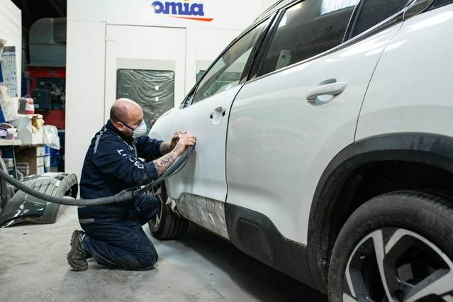 Dans un atelier de réparation, un mécanicien est agenouillé en train de poncer une portière de voiture blanche.